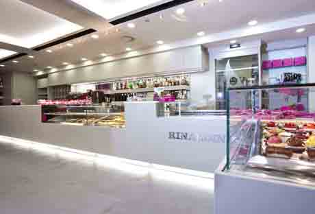 Interno pasticceria Rinaldini: bancone bianco illuminato, vetrine con dolci e torte. Design moderno e luminoso.