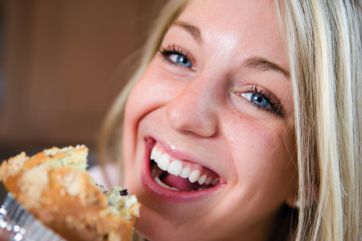 Donna bionda sorride mentre mangia un muffin. Occhi azzurri, felicità, colazione.