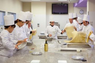 Corso di pasticceria: studenti ALMA preparano pasta fresca. Divise bianche e cappelli da chef. Cucina professionale.