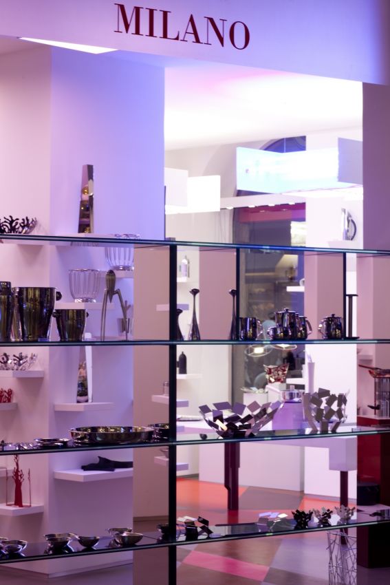 Vetrina negozio Milano: oggetti di design in vetro e metallo su scaffali illuminati. Arredamento moderno e lussuoso.