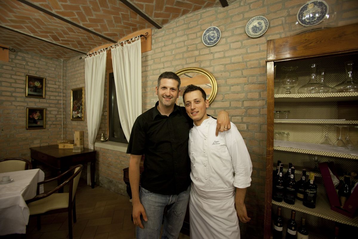 Ristorante italiano: chef e cliente sorridono. Interni rustici, mattoni a vista, arredamento classico. Cucina tipica.