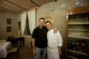 Ristorante italiano: chef e cliente sorridono. Interni rustici, mattoni a vista, arredamento classico. Cucina tipica.