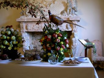 Tavolo imbandito con frutta, fiori e fagiano. Camino decorativo sullo sfondo. Decorazioni autunnali.