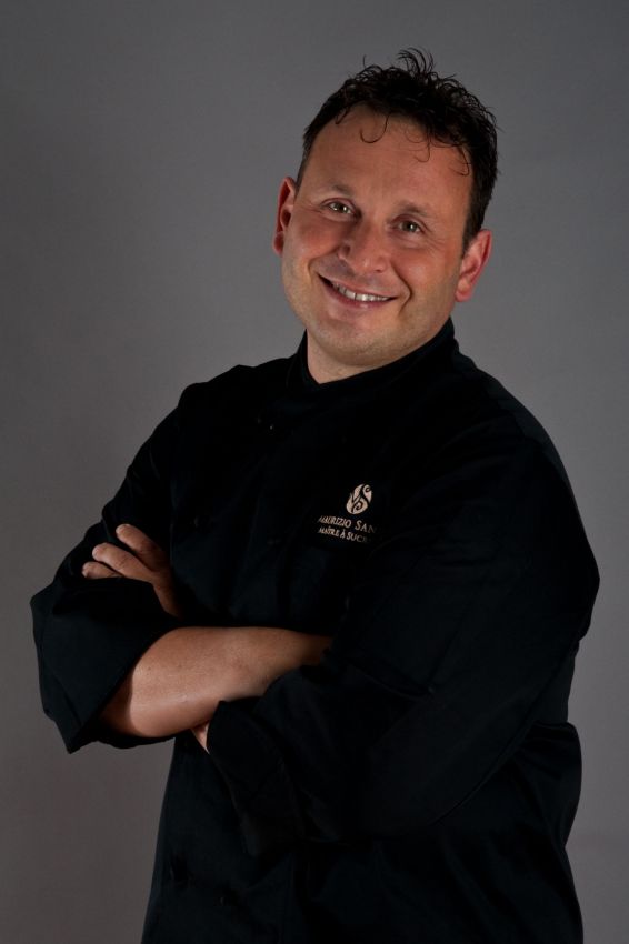 Ritratto di Maurizio Santin, chef in divisa nera, braccia conserte e sorriso. Cucina italiana di alta qualità.