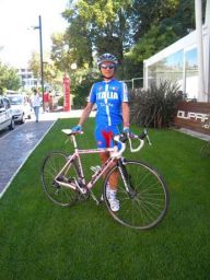 Ciclista italiana con bici da corsa rosa su prato. Abbigliamento sportivo azzurro. Casco e occhiali da sole.