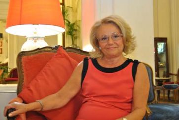 Ritratto di Fausta, signora elegante seduta su poltrona rossa. Occhiali, vestito corallo.