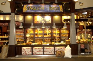 Panificio artigianale: scaffali pieni di pane fresco, sacchi di farina e insegna Täglich Brot. Panificazione tradizionale.