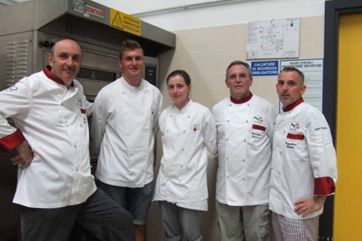 Squadra italiana di chef in cucina. Divise bianche, colli rossi. Forno professionale Zanoli sullo sfondo.
