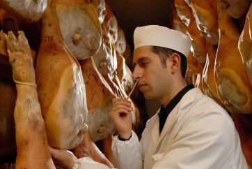 Produzione Prosciutto San Daniele: esperto controlla stagionatura con ago. Salumi DOP appesi.