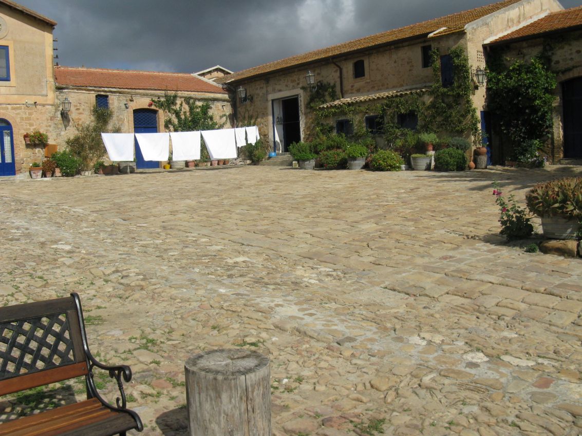 Cortile in pietra di masseria siciliana con panni stesi, panchina e cielo nuvoloso. Architettura rurale, Sicilia.