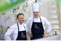 Due chef sorridenti con grembiuli neri. Cucina Tschuggen Grand Hotel Arosa.