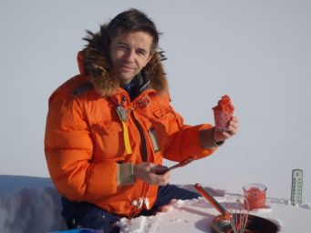 Uomo in giacca arancione prepara cibo rosso sulla neve. Cucina invernale, avventura culinaria.