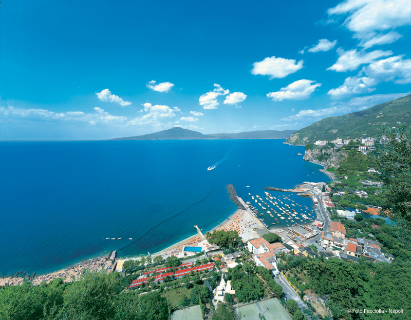 Panorama mozzafiato Le Axidie: mare cristallino, Vesuvio sullo sfondo, costa frastagliata e porticciolo turistico.