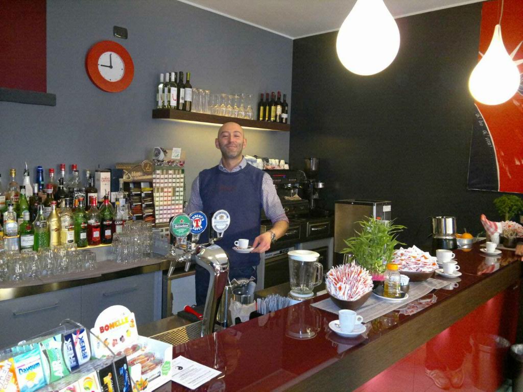 Barista sorridente al bar. Bottiglie di alcolici, caffè, lampade pendenti. Arredamento bar italiano.