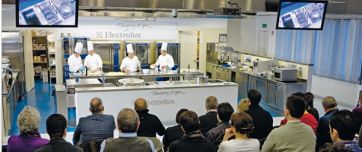 Corso di cucina Electrolux: chef al lavoro, pubblico attento. Cucina professionale, elettrodomestici inox.