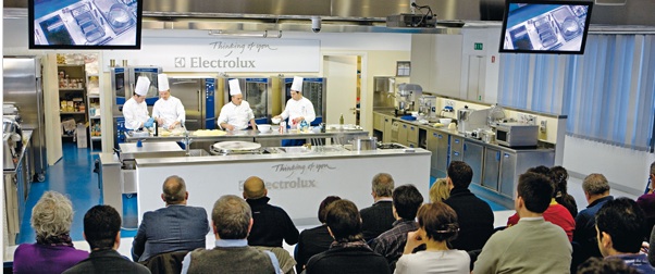 Corso di cucina Electrolux: chef al lavoro, pubblico attento. Cucina professionale, elettrodomestici inox.