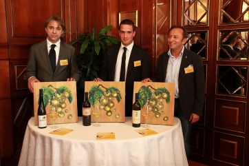 Degustazione vini italiani: tre uomini presentano bottiglie e quadri di grappoli d'uva su tavolo rotondo.