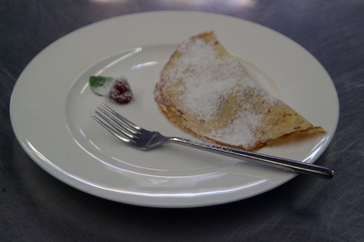 Crêpe dolce con zucchero a velo su piatto bianco. Forchetta e lampone come decorazione.