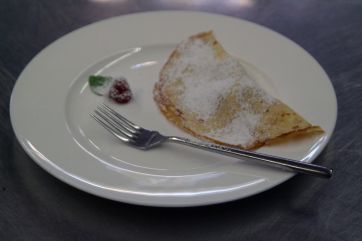 Crêpe dolce con zucchero a velo su piatto bianco. Forchetta e lampone come decorazione.