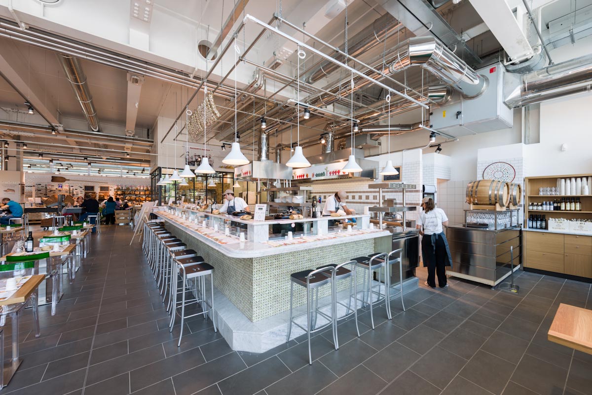 Interno di Eataly: bar con sgabelli, illuminazione industriale e cucina a vista. Ristorante italiano.
