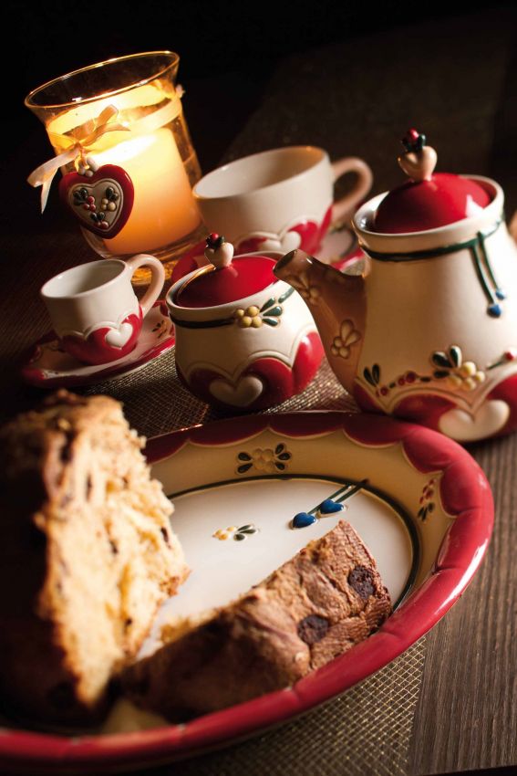 Natura morta: teiera, tazza, panettone. Ceramiche decorate, candela accesa. Atmosfera calda e accogliente.