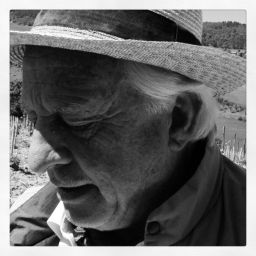 Ritratto in bianco e nero di uomo anziano con cappello. Dettaglio del volto segnato dal tempo. Campagna sullo sfondo.