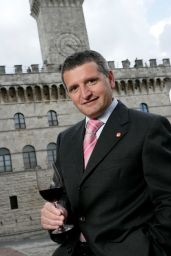 Uomo in giacca e cravatta con bicchiere di vino rosso. Torre medievale sullo sfondo.