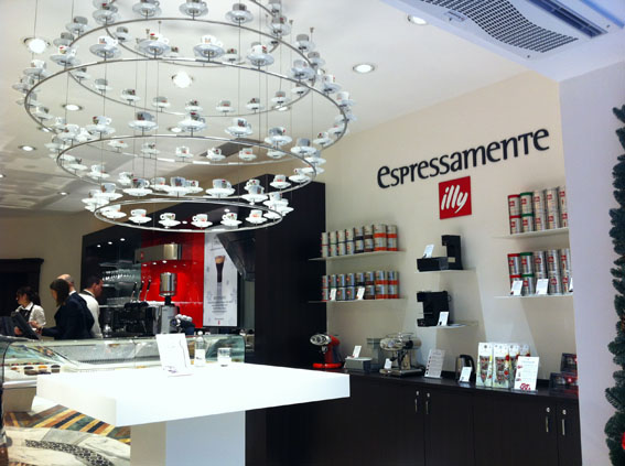 Interno di caffetteria Espressamente Illy: lampadario di tazze, scaffali con caffè Illy.