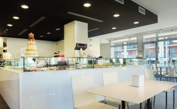Interno di pasticceria moderna con vetrina dolci, tavolo bianco e sedie. Design minimalista e luminoso.