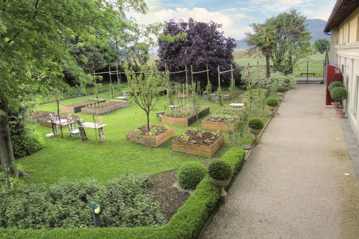 Giardino curato con aiuole rialzate in legno, prato verde e vialetto di ghiaia. Arredi rustici e piante ornamentali.