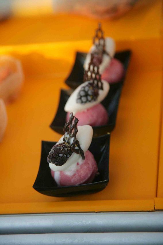 Finger food dolce: palline gelato rosa, crema bianca e decorazioni cioccolato su cucchiai neri.