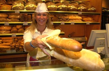 Panettiera sorridente offre pane fresco. Scaffali pieni di pane artigianale in panetteria.