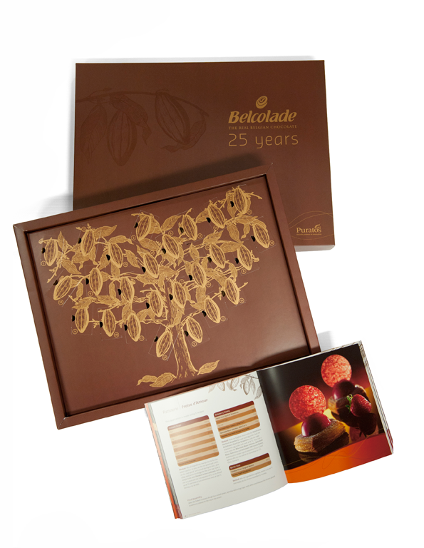 Scatola Belcolade 25 anni: cioccolato belga artigianale. Design elegante con albero di cacao dorato. Ricettario incluso.