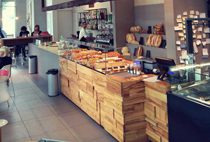 Interno di bar/caffetteria con bancone in legno, esposizione prodotti da forno e sedute per clienti.