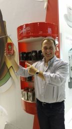 Uomo sorridente mostra caffè Lucaffè. Scaffale rosso con barattoli di caffè. Marchio italiano, alta qualità.