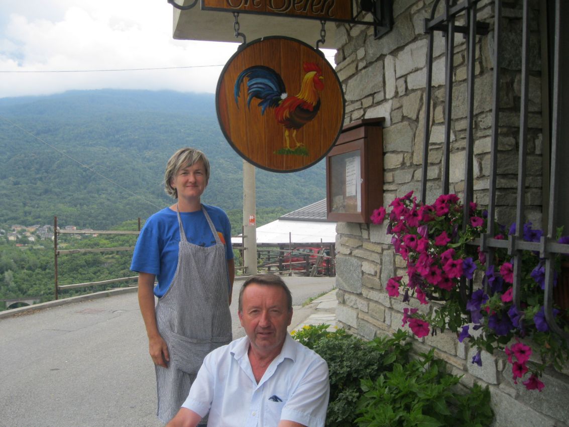 Gianluca e Serena Martina davanti al ristorante. Insegna con gallo, fiori e vista sulle montagne.