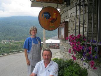 Gianluca e Serena Martina davanti al ristorante. Insegna con gallo, fiori e vista sulle montagne.