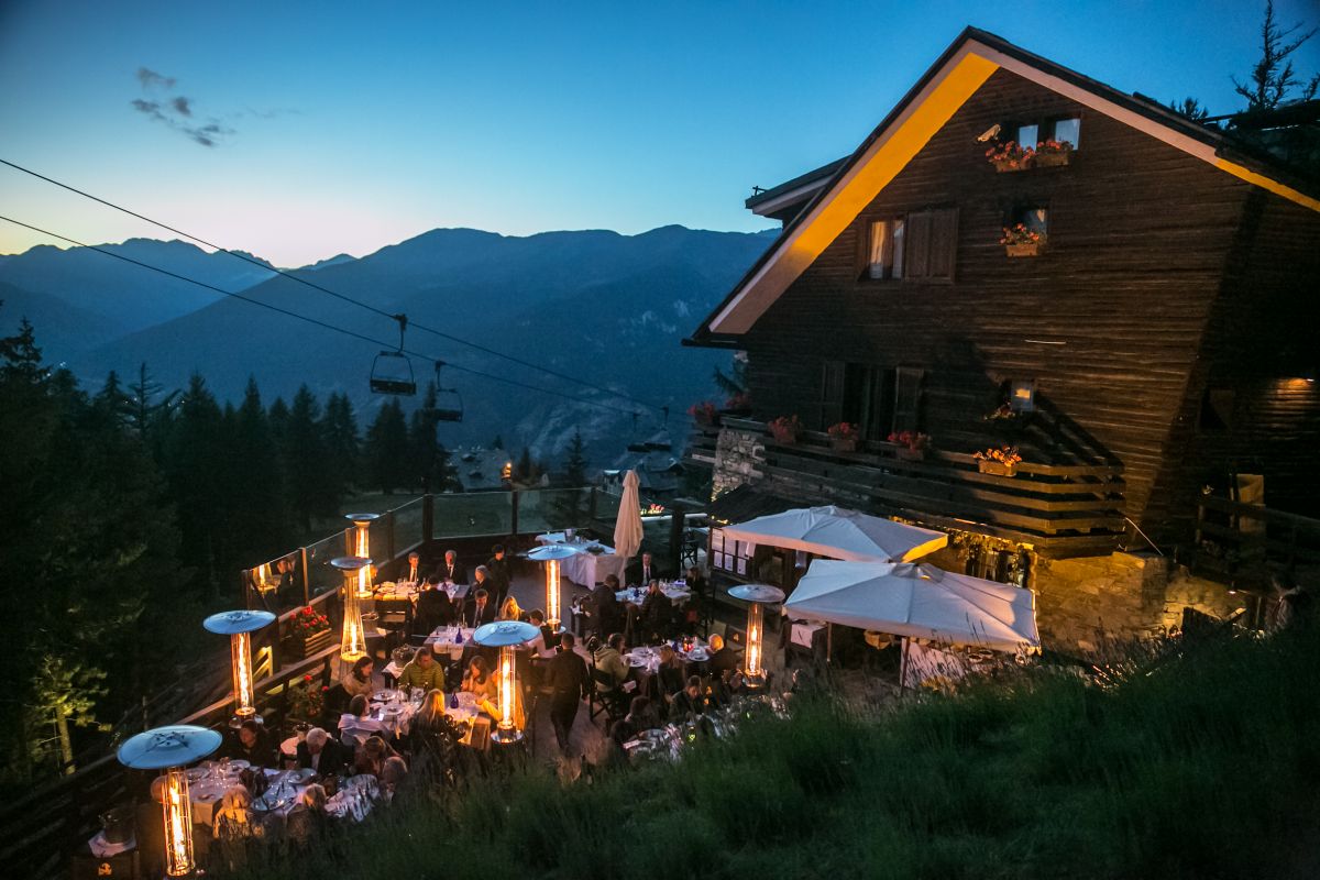 Cena elegante in chalet alpino. Tavoli imbanditi, riscaldatori, vista montagne e funivia al tramonto.