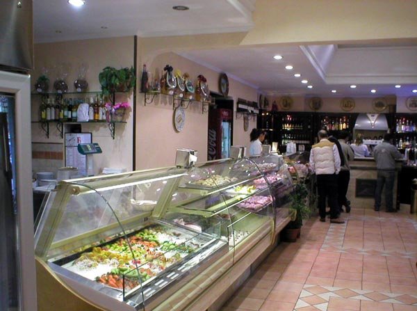 Gastronomia italiana: vetrina con insalate fresche e piatti pronti. Clienti al bancone bar. Bottiglie sullo scaffale.