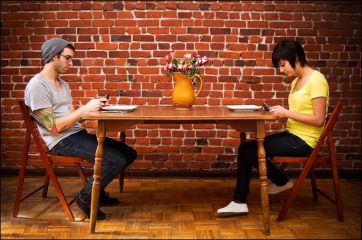 Coppia al ristorante, appuntamento. Lui con tatuaggio e cappello, lei con maglia gialla, entrambi usano il cellulare. Muro di mattoni.