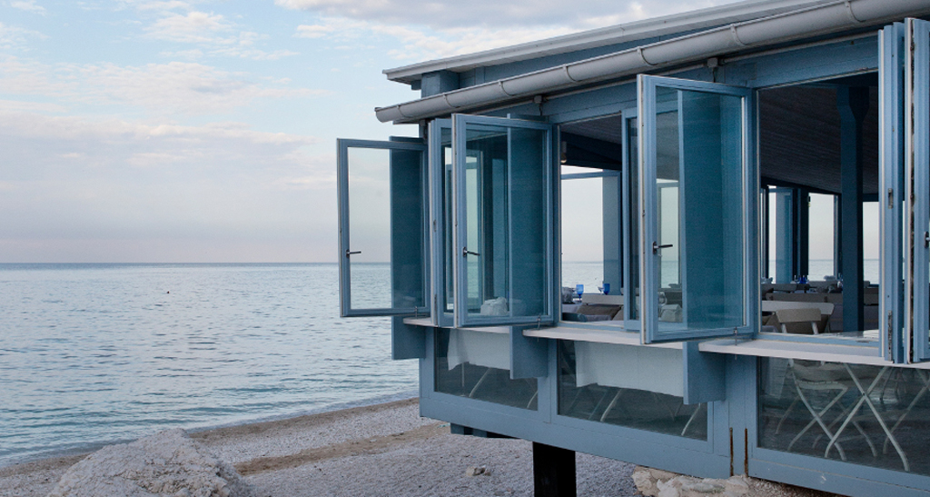 Ristorante sul mare con finestre aperte. Vista sull'Adriatico, spiaggia di sassi. Design costiero.