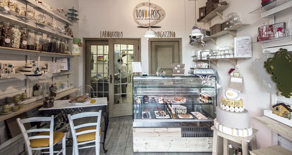 Interno di Uovo a Pois, pasticceria artigianale. Vetrina dolci, scaffali con biscotti e decorazioni. Design accogliente.