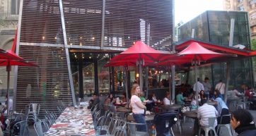 Ristorante all'aperto con tavoli, sedie e ombrelloni rossi. Persone sedute e atmosfera vivace.