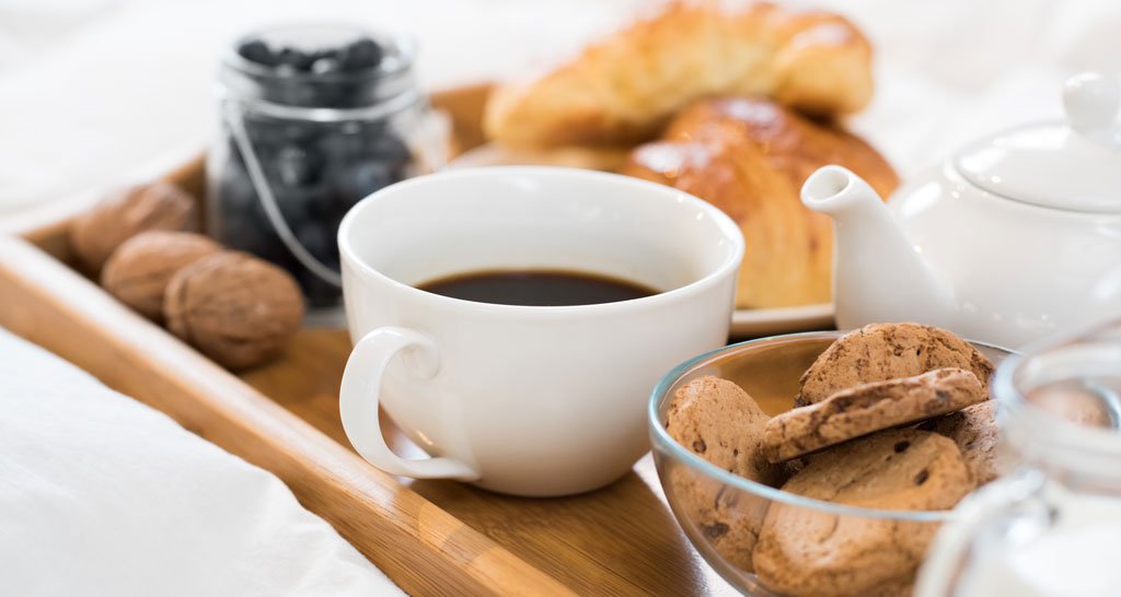 Colazione a letto: caffè, croissant, biscotti, mirtilli e noci su vassoio di legno.