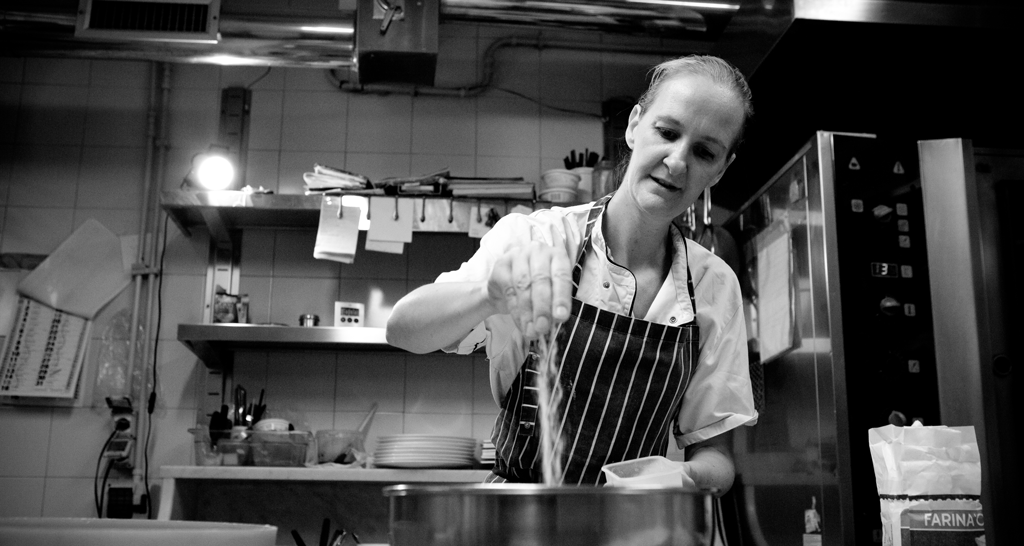 Chef in cucina. Donna con grembiule aggiunge farina. Ristorante in bianco e nero.