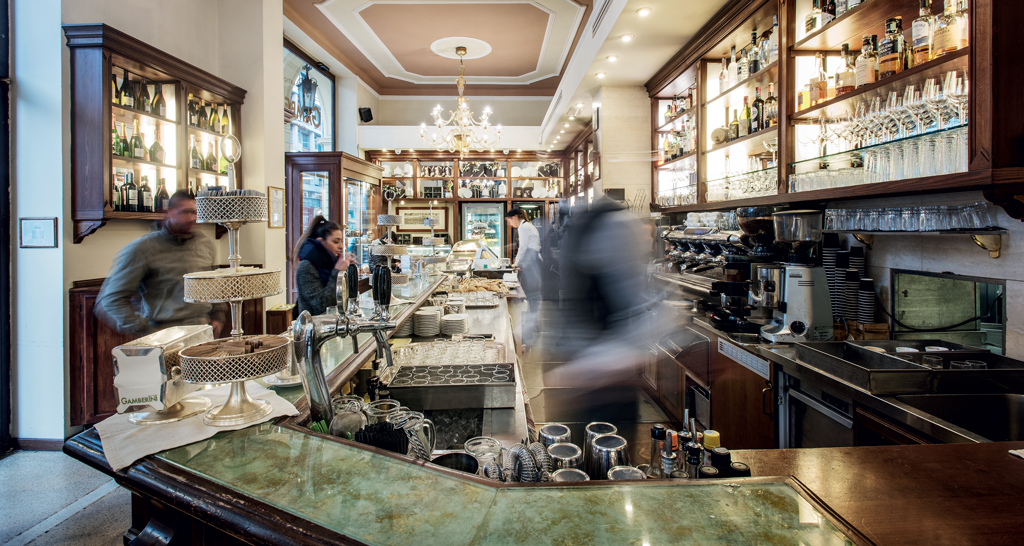 Interno del Caffè Gamberini a Bologna. Bancone bar, lampadario, scaffali con bottiglie e persone.