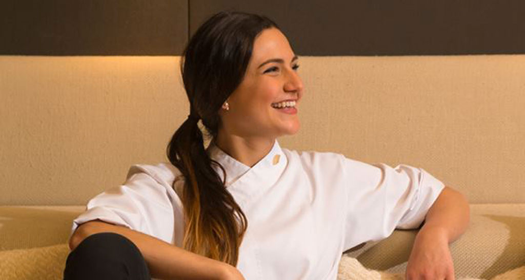 Chef Andrea Dopico Cafarelli sorride. Divisa bianca da cucina, capelli scuri raccolti.