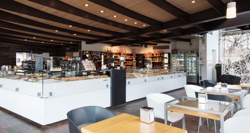 Interno di pasticceria: vetrine con dolci, tavoli e sedie. Arredamento moderno e luminoso.