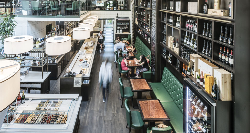 Interno di bottega con scaffali di vino, tavoli e sedute verdi. Illuminazione moderna e atmosfera accogliente.