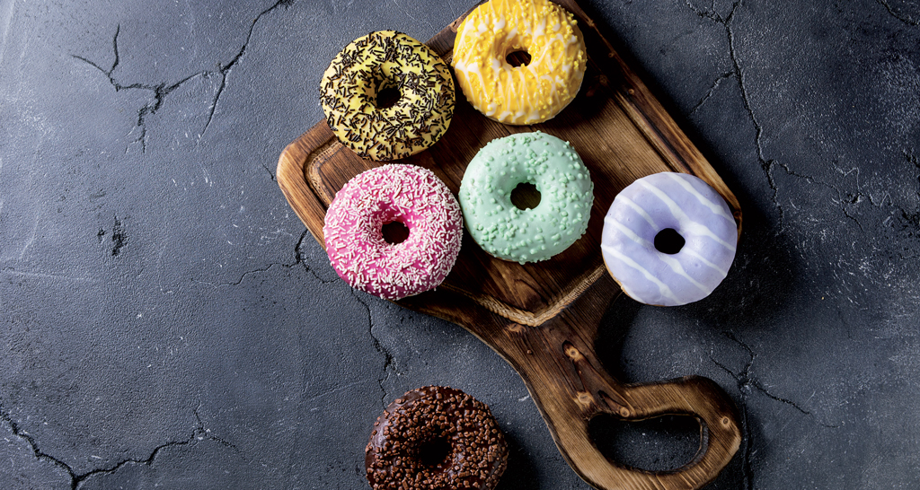 Ciambelle colorate su tagliere. Dolci glassati assortiti: rosa, giallo, verde, viola e cioccolato. Dessert golosi.